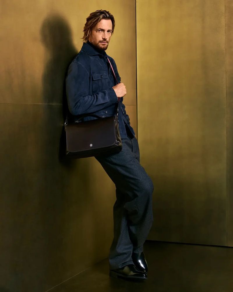 Full-length shot of an adult man leaning against a gold wall wearing a navy jacket, wide leg jeans, and black leather messenger bag.