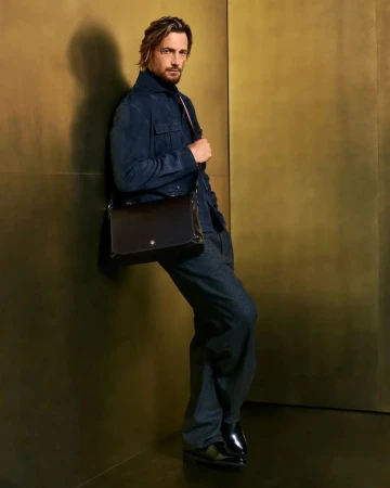 Full-length shot of an adult man leaning against a gold wall wearing a navy jacket, wide leg jeans, and black leather messenger bag.