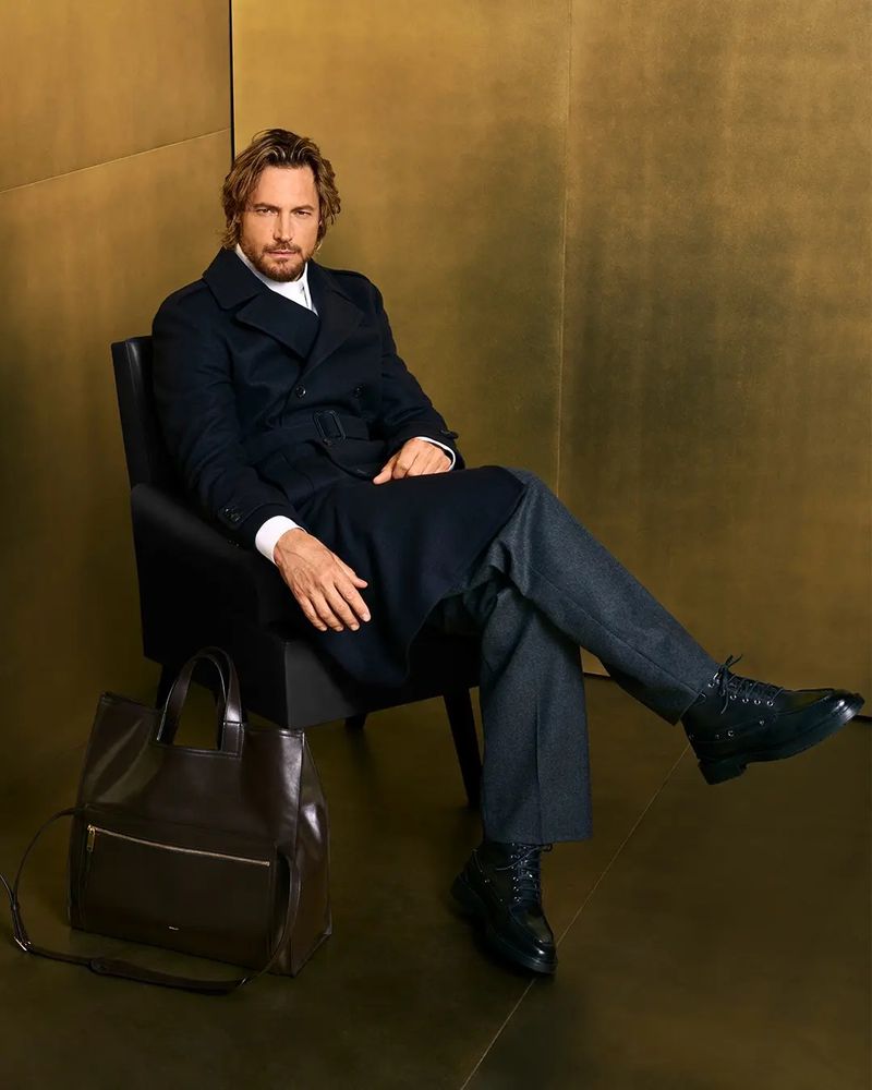 Adult male with blond hair sits in black chair wearing dark coat, grey pants, and black boots, next to dark brown leather bag.