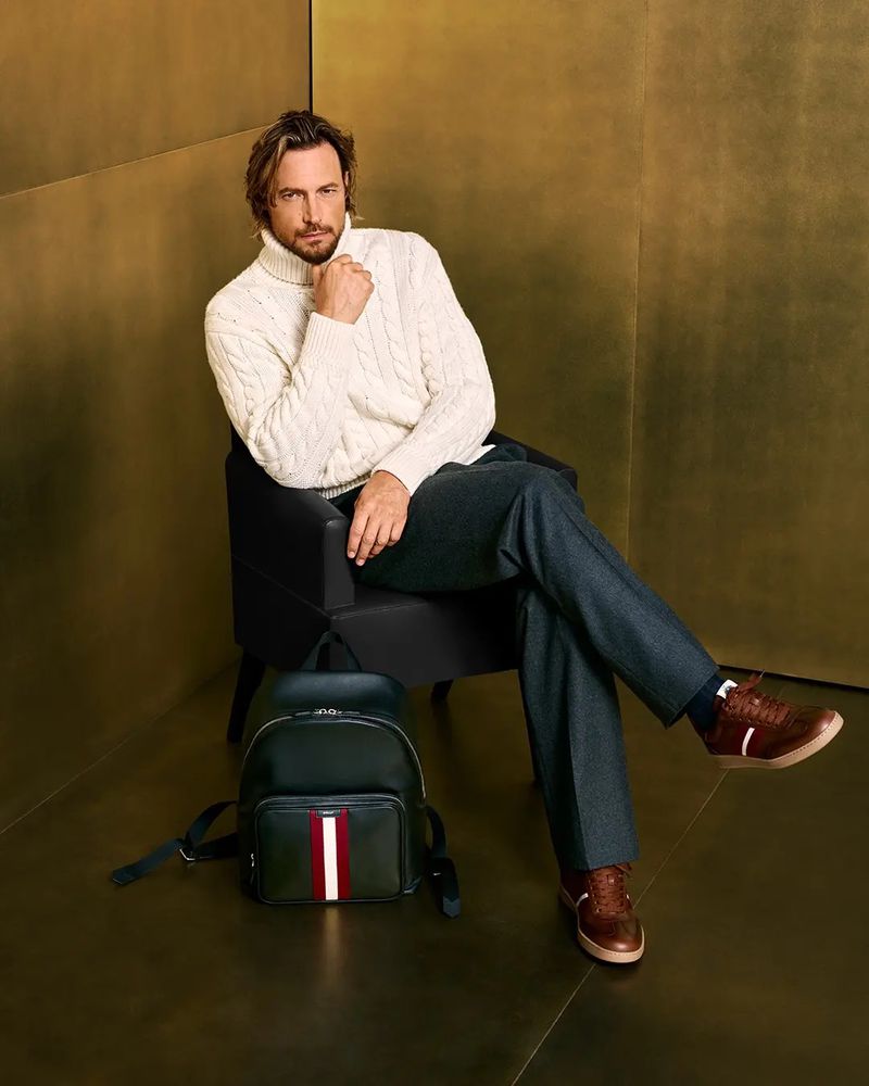 Male model in cream turtleneck sweater seated in black chair next to backpack and wearing brown leather shoes against gold backdrop.