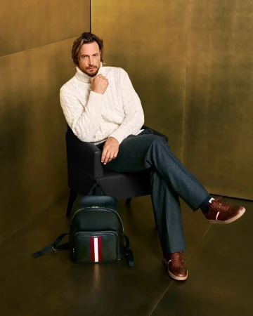 Male model in cream turtleneck sweater seated in black chair next to backpack and wearing brown leather shoes against gold backdrop.