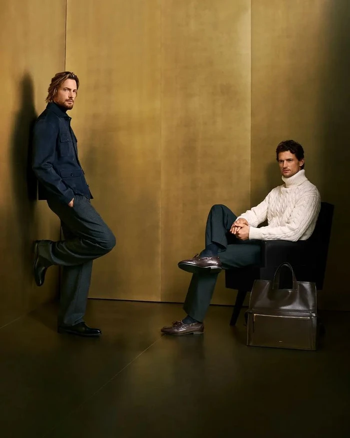 Two men posing in a luxury campaign by Bally, featuring a standing man in a dark jacket and a seated man in a white turtleneck.