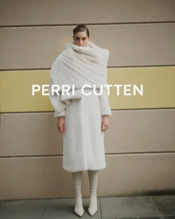 Fashion shot of a woman wearing a white coat and matching boots standing in front of a tan and yellow wall with the text PERRI CUTTEN.