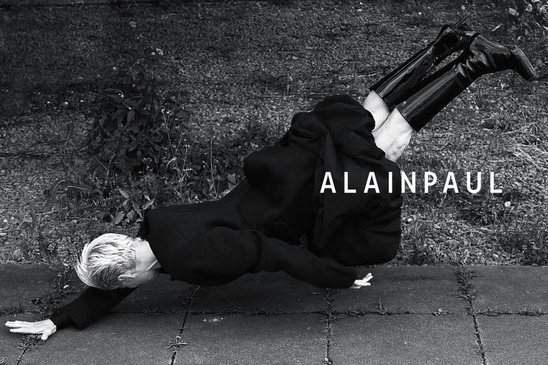 Editorial fashion photo of an adult woman in a black coat and boots doing a handstand on a stone walkway outdoors, shot in black and white.