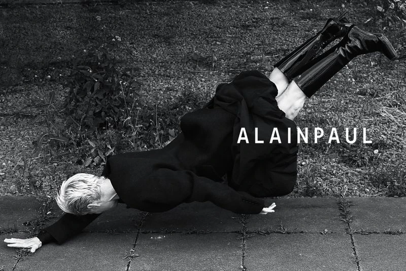 Editorial fashion photo of an adult woman in a black coat and boots doing a handstand on a stone walkway outdoors, shot in black and white.