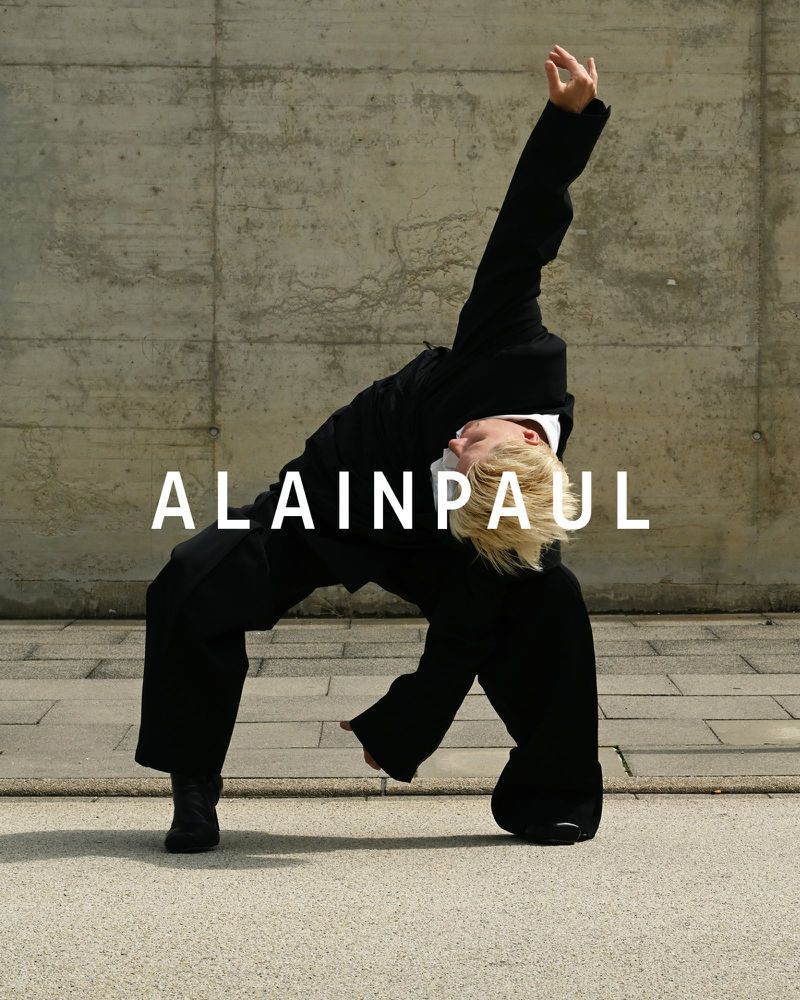 Person in a black suit bends backward in an open, urban setting against a textured concrete wall, showcasing a modern, avant-garde style.