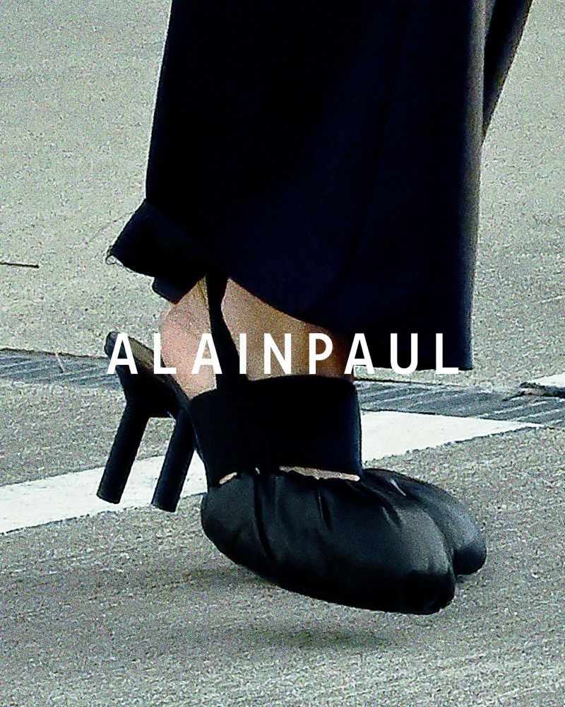 Close-up on a pair of black, high-heeled shoes with a draped, ruched toe, set against a neutral grey ground with drain or expansion joints.