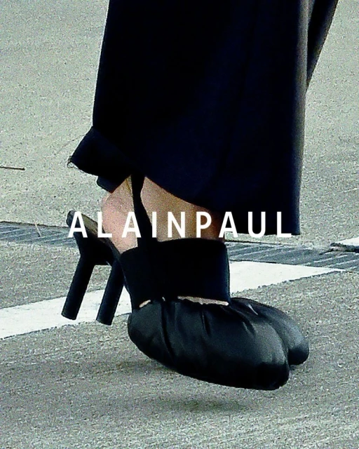 Close-up on a pair of black, high-heeled shoes with a draped, ruched toe, set against a neutral grey ground with drain or expansion joints.