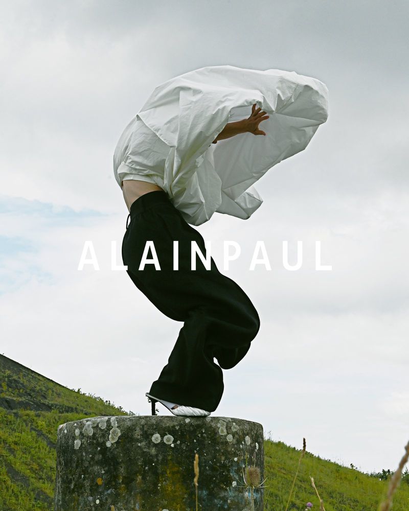 A person in black pants and silver heels stands on a concrete column, their head covered by a white sheet in a field under a cloudy sky.