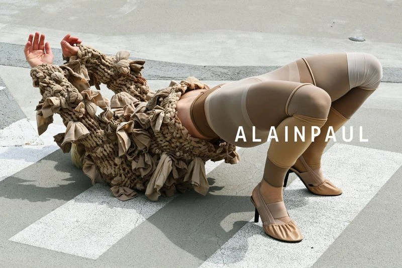 Fashion shot: a person in artistic, beige ensemble strikes a pose on a painted crosswalk, paired with beige heels, set against a concrete backdrop.