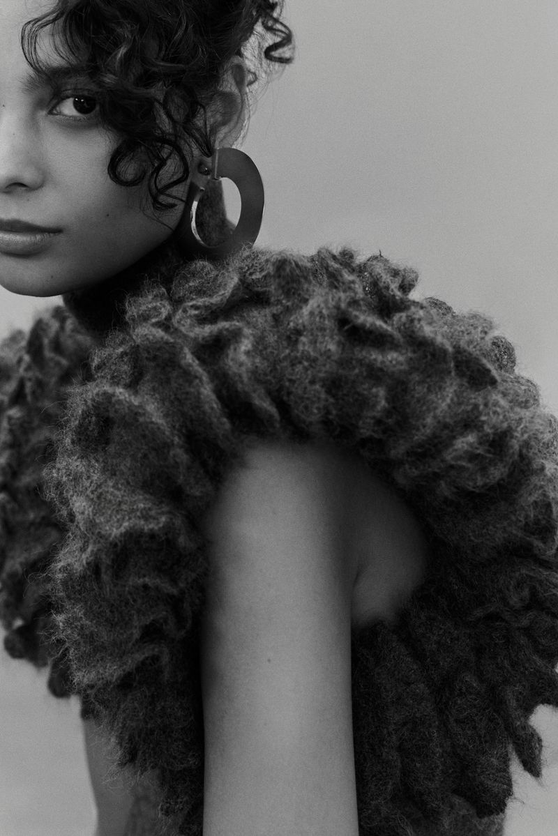 Close-up portrait of a curly-haired person with dark hair and an earring posing in front of a plain grey backdrop, wearing a fuzzy collar.