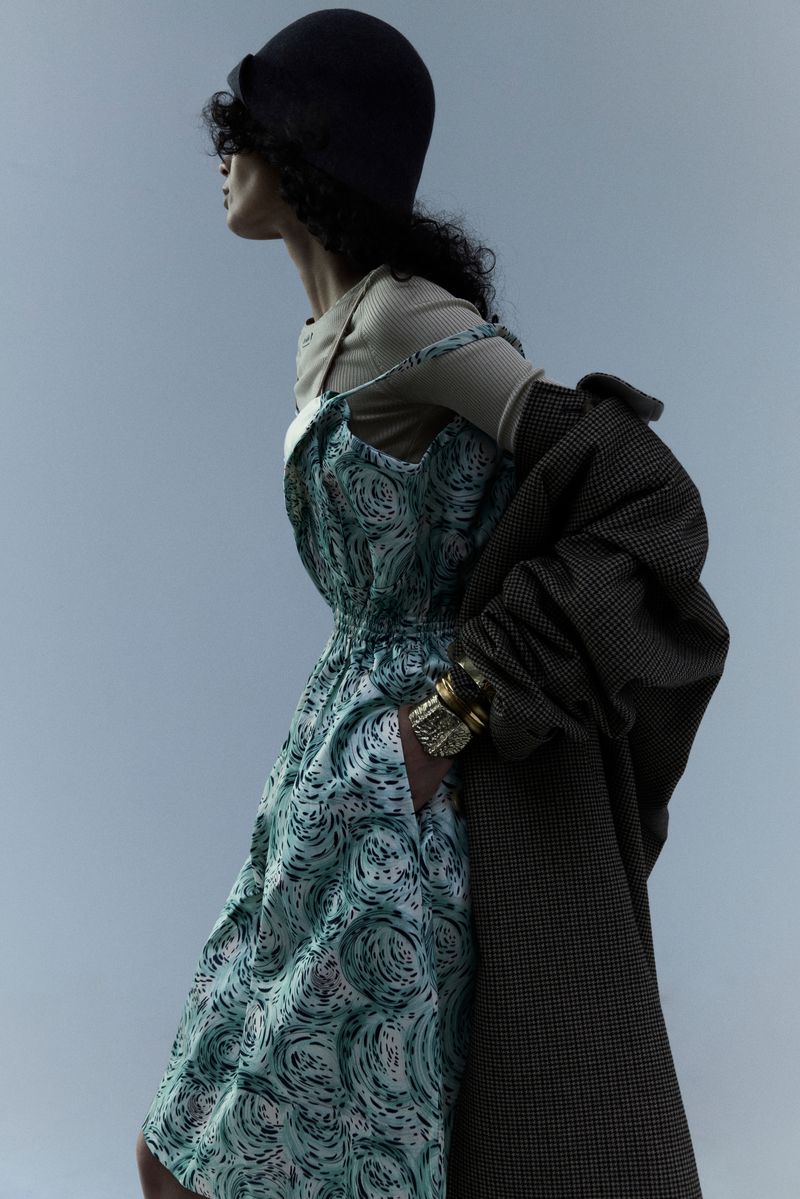 A person wears a patterned dress and dark coat in a studio shot, against a light blue background, accessorized with a dark hat and gold cuffs.