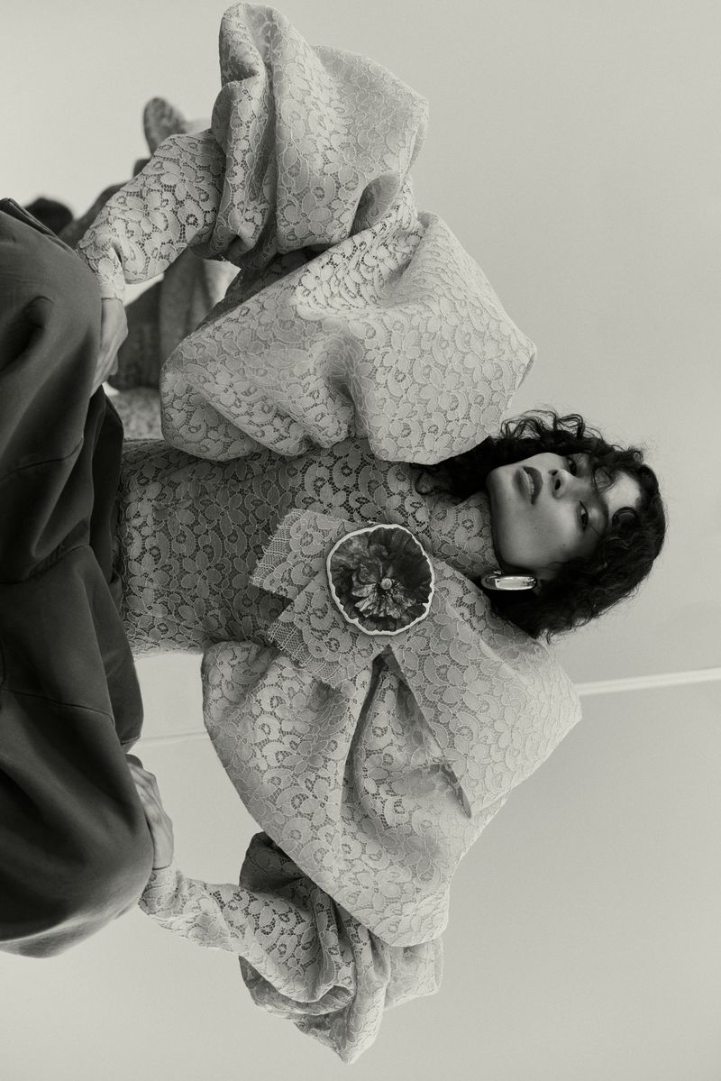 Monochromatic fashion shot featuring a single adult female model in an ornate, vintage-inspired dress with large sleeves and a floral brooch.
