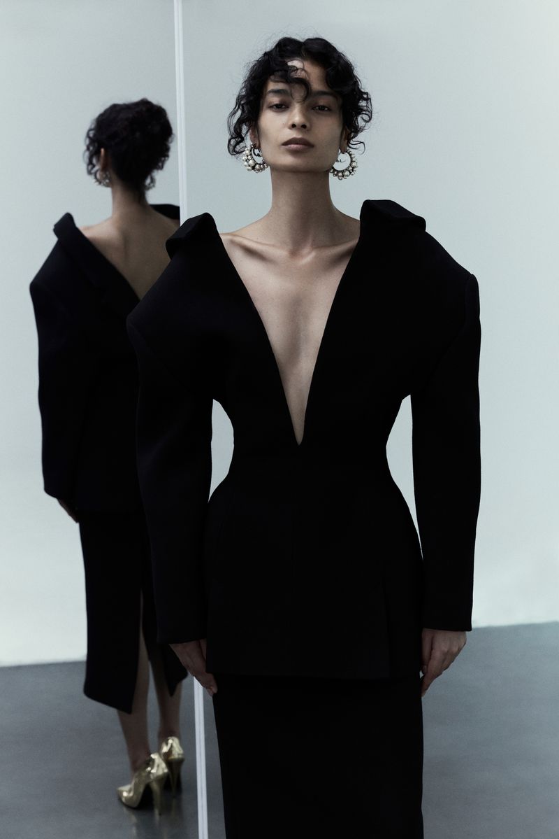 Elegant woman in a black sculptural dress poses in a photo studio next to a large mirror reflecting her back, fashion portrait.