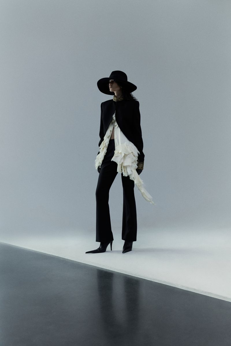 Fashion model poses in a black blazer, wide-leg pants, and wide-brimmed hat against a neutral backdrop in a studio fashion shot.