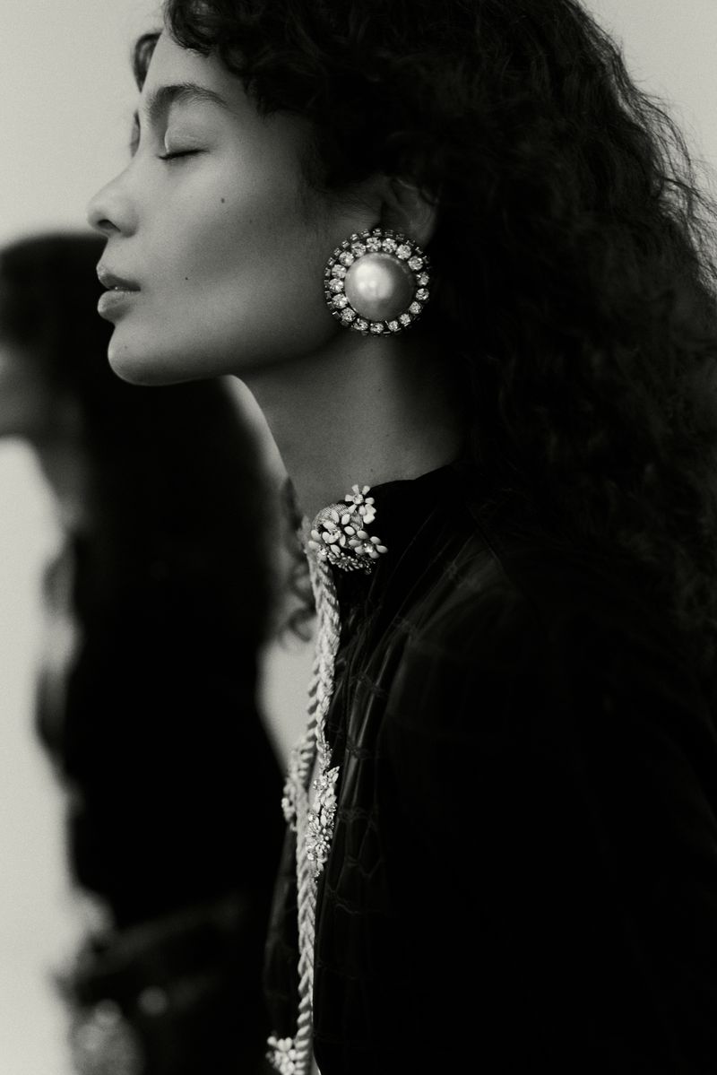 Monochrome portrait of a woman with eyes closed, showcasing pearl earrings, a high-necked embellished top, and soft, curly hair.