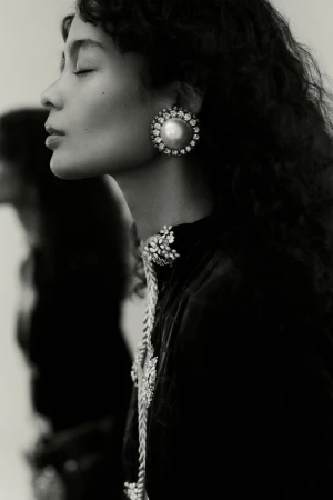 Monochrome portrait of a woman with eyes closed, showcasing pearl earrings, a high-necked embellished top, and soft, curly hair.