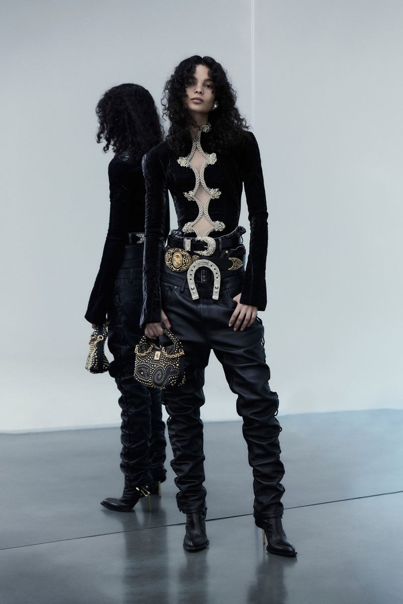 A woman poses in a black outfit with horseshoe-detailed belt, holding a beaded bag, with a mirror reflecting her image in a light studio.