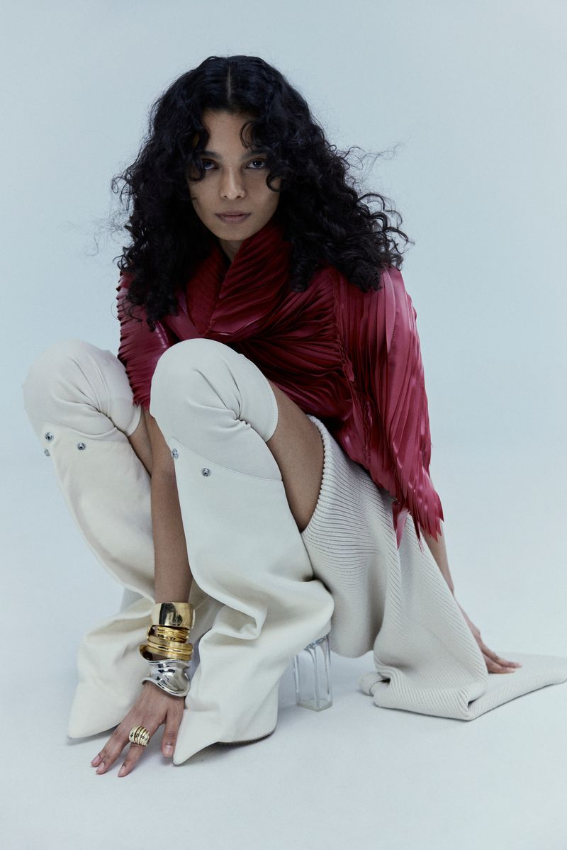 Fashionable woman with curly hair poses in an indoor studio with bright lighting, wearing a red top and futuristic white boots.