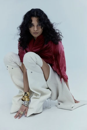 Fashionable woman with curly hair poses in an indoor studio with bright lighting, wearing a red top and futuristic white boots.