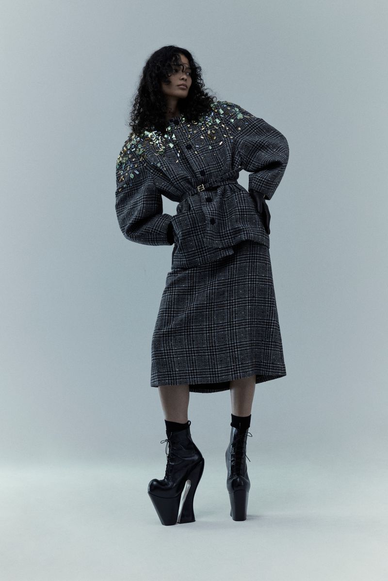 A woman in a plaid suit and platform boots poses against a light gray backdrop, showcasing avant-garde fashion with a unique silhouette.