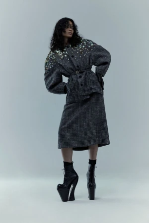 A woman in a plaid suit and platform boots poses against a light gray backdrop, showcasing avant-garde fashion with a unique silhouette.