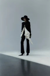 Full-length portrait of a model wearing a wide-brimmed black hat, structured jacket, and flowing ruffled white top against a grey studio backdrop.