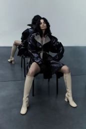 Model seated in a black coat and tall beige boots with a mirror reflection