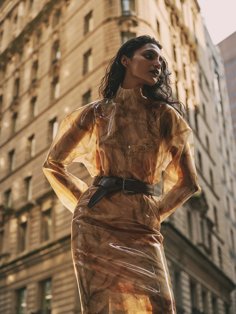 A young adult woman poses in a translucent brown dress with a wide black belt against a high-rise urban building in the daytime.