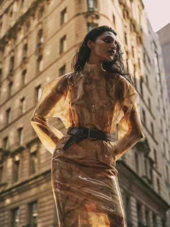 A young adult woman poses in a translucent brown dress with a wide black belt against a high-rise urban building in the daytime.