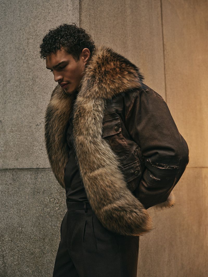 Portrait of a stylish adult male in a brown fur-collared leather jacket and black trousers, standing against a textured wall.
