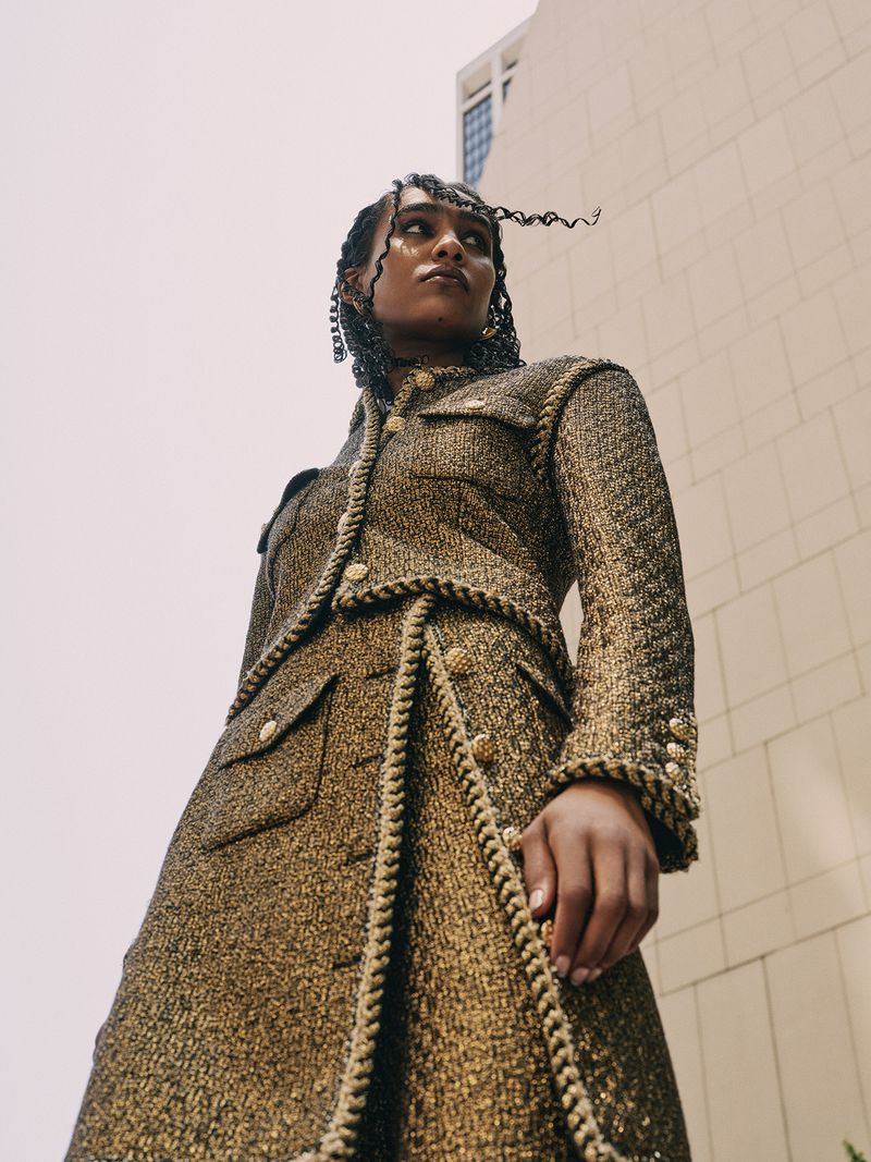 Low-angle shot of an adult female in a golden tweed skirt suit, posing outdoors against a tall building with a cloudy sky backdrop.