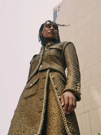 Low-angle shot of an adult female in a golden tweed skirt suit, posing outdoors against a tall building with a cloudy sky backdrop.