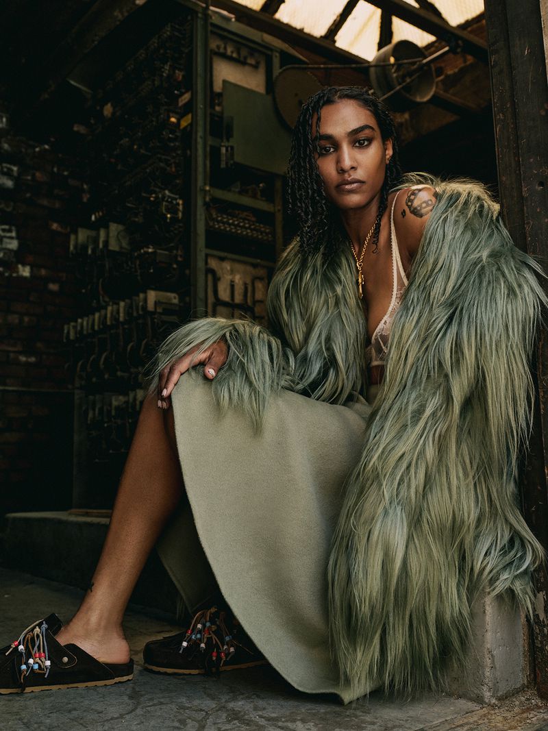 Indoor fashion shot of a woman with dark hair wearing a light green coat, a skirt, and dark clogs with beaded accents.