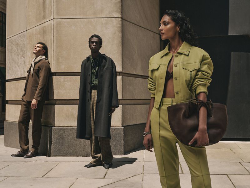 Fashion editorial shot of three models outdoors, wearing coordinated modern clothing and accessories, standing on an urban street setting.