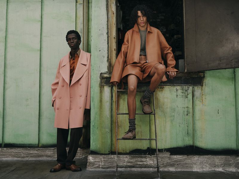 Two male models in stylish earth-toned outfits pose outdoors against a textured, light green wall with industrial accents in a fashion editorial.