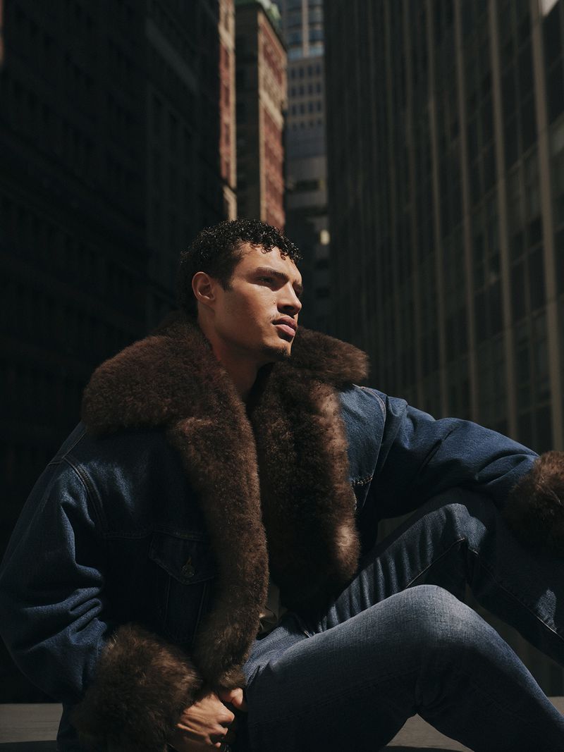 Male model sits in a city environment, wearing a denim jacket with fur trim, denim pants, bathed in natural light with dark shadows.