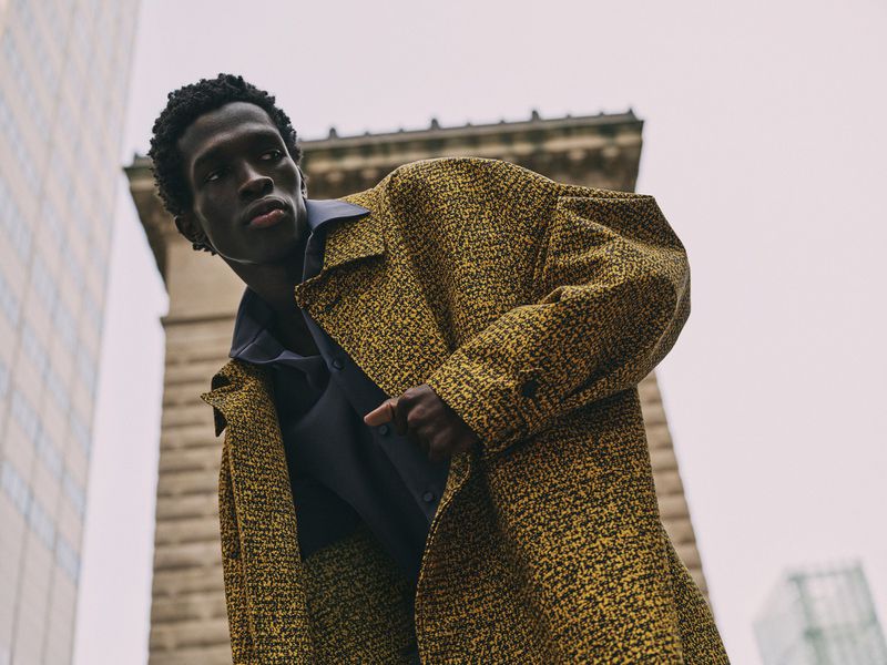 Low-angle shot of a Black male model in a yellow tweed coat in an urban setting, with skyscrapers in the background, capturing fashion and style.