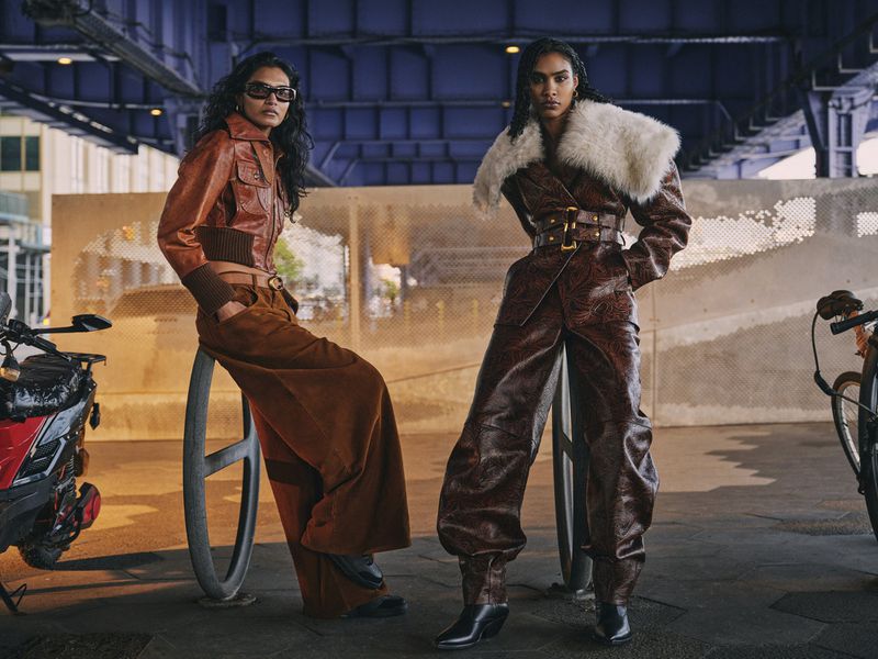 Two women in stylish leather outfits pose outdoors under an overpass with bicycles and a scooter, showcasing urban fashion.