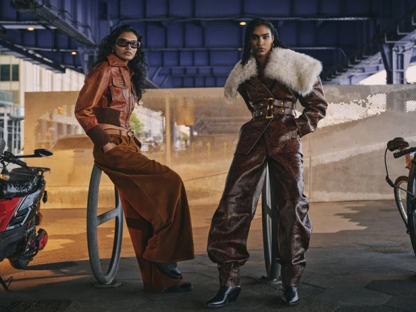 Two women in stylish leather outfits pose outdoors under an overpass with bicycles and a scooter, showcasing urban fashion.