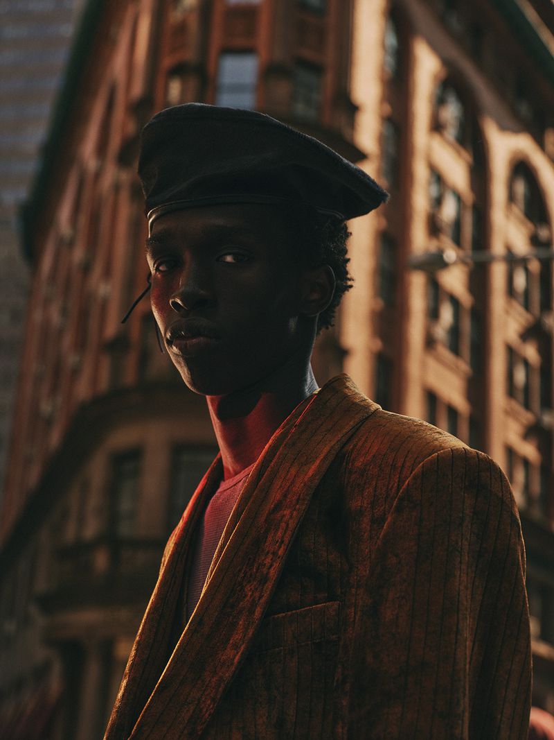 Portrait of a black adult male wearing a beret and blazer, standing in an urban environment with buildings in the background.