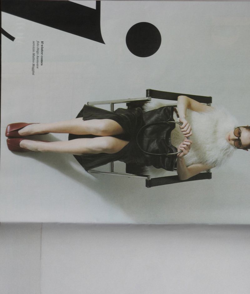 Fashion editorial shot of a woman lying in a chrome chair with a white fur collar, dark dress, maroon shoes, and sunglasses in a studio.
