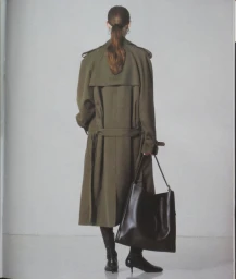 Model seen from behind wearing an olive green trench coat and holding a large black leather shoulder bag against a grey seamless backdrop
