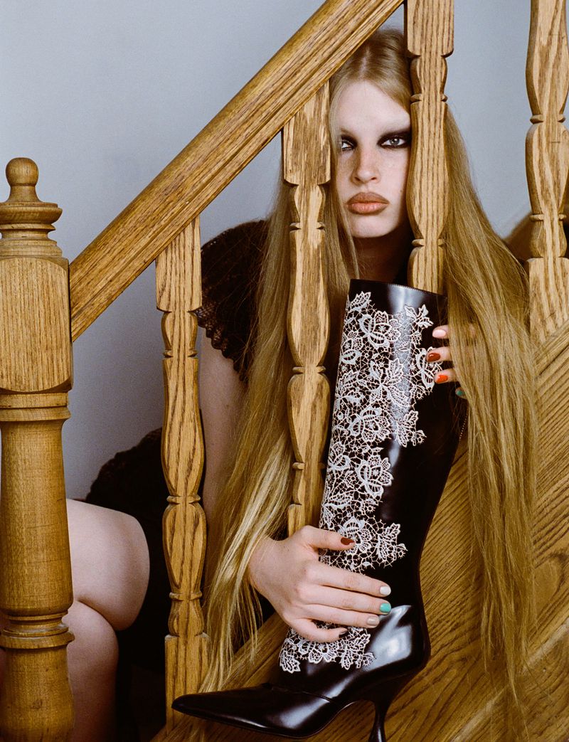 Fashion editorial shot of a model holding a high-heeled boot with a lace design while seated on a wood staircase indoors.