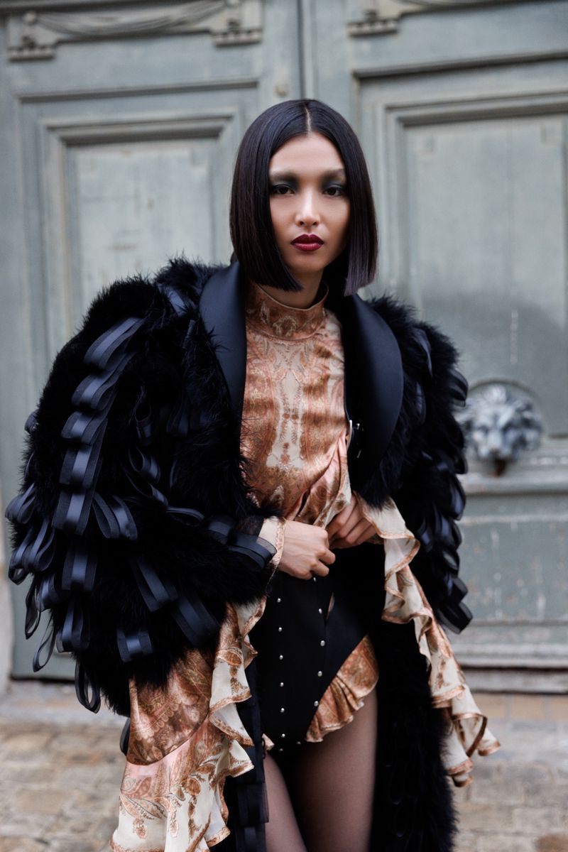 Fashionable woman poses outdoors wearing a feather and fur coat over a paisley print mini-dress in front of a door.