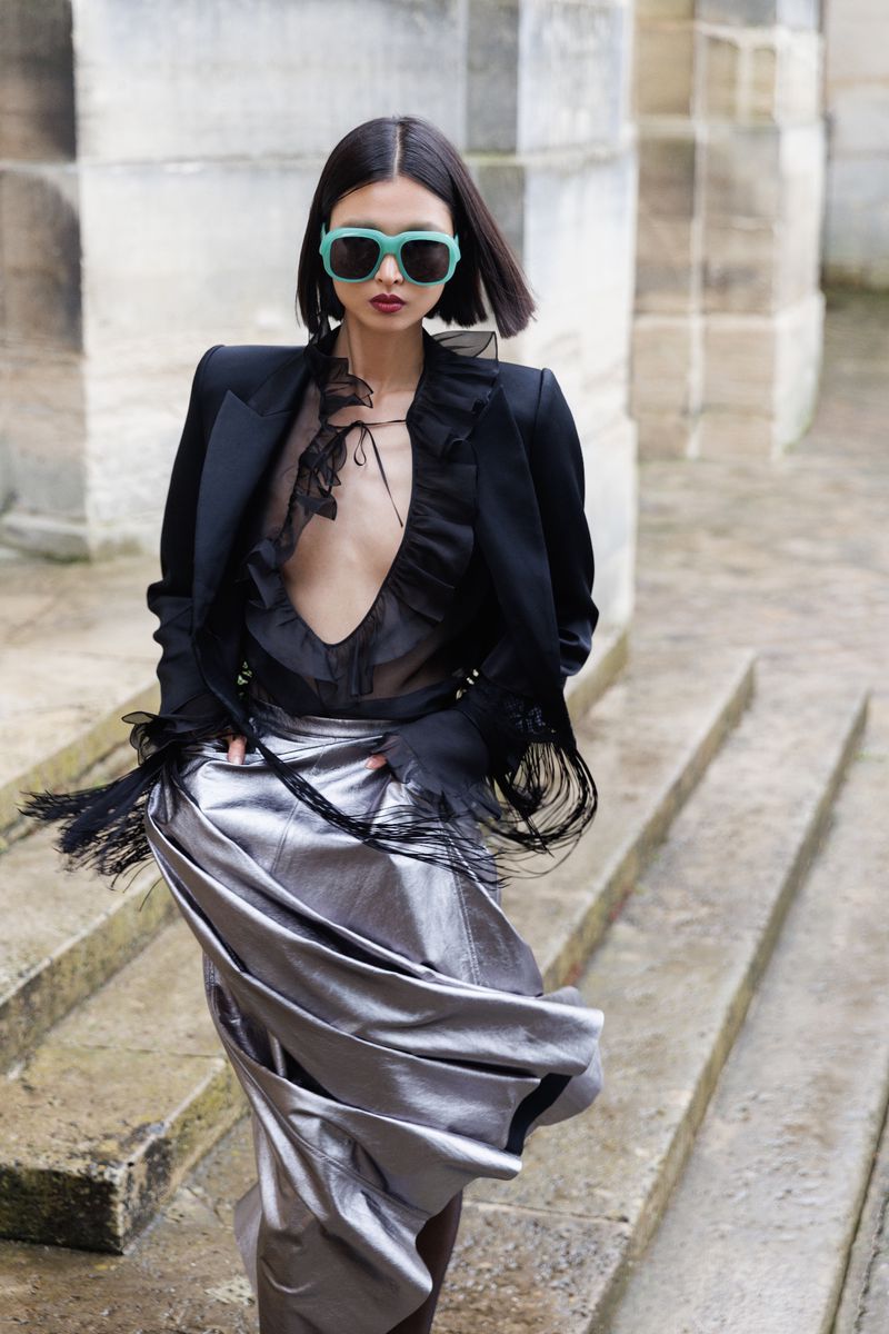 A stylish woman wearing a black blazer, sheer black blouse, and metallic silver skirt poses on stone steps with a blurred background.