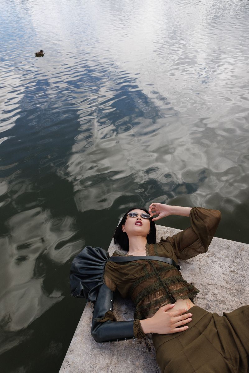 Woman in olive green outfit and sunglasses lies on a stone platform next to a pond reflecting the sky on a cloudy day.