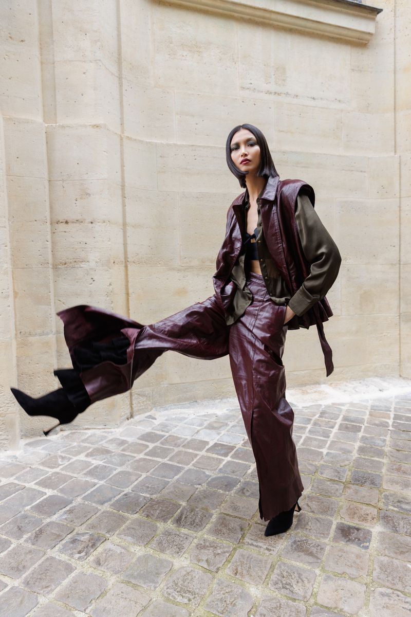A fashionable adult woman wearing leather-like pants and vest poses on a stone path outdoors with architectural details.