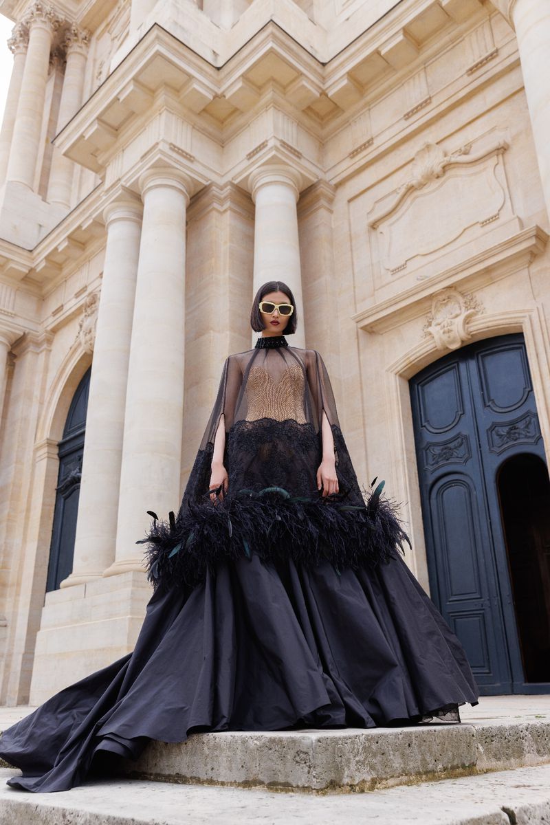 Elegant woman in a black feathered gown stands on stone steps in front of a grand, light-stone building with columns and arched windows.