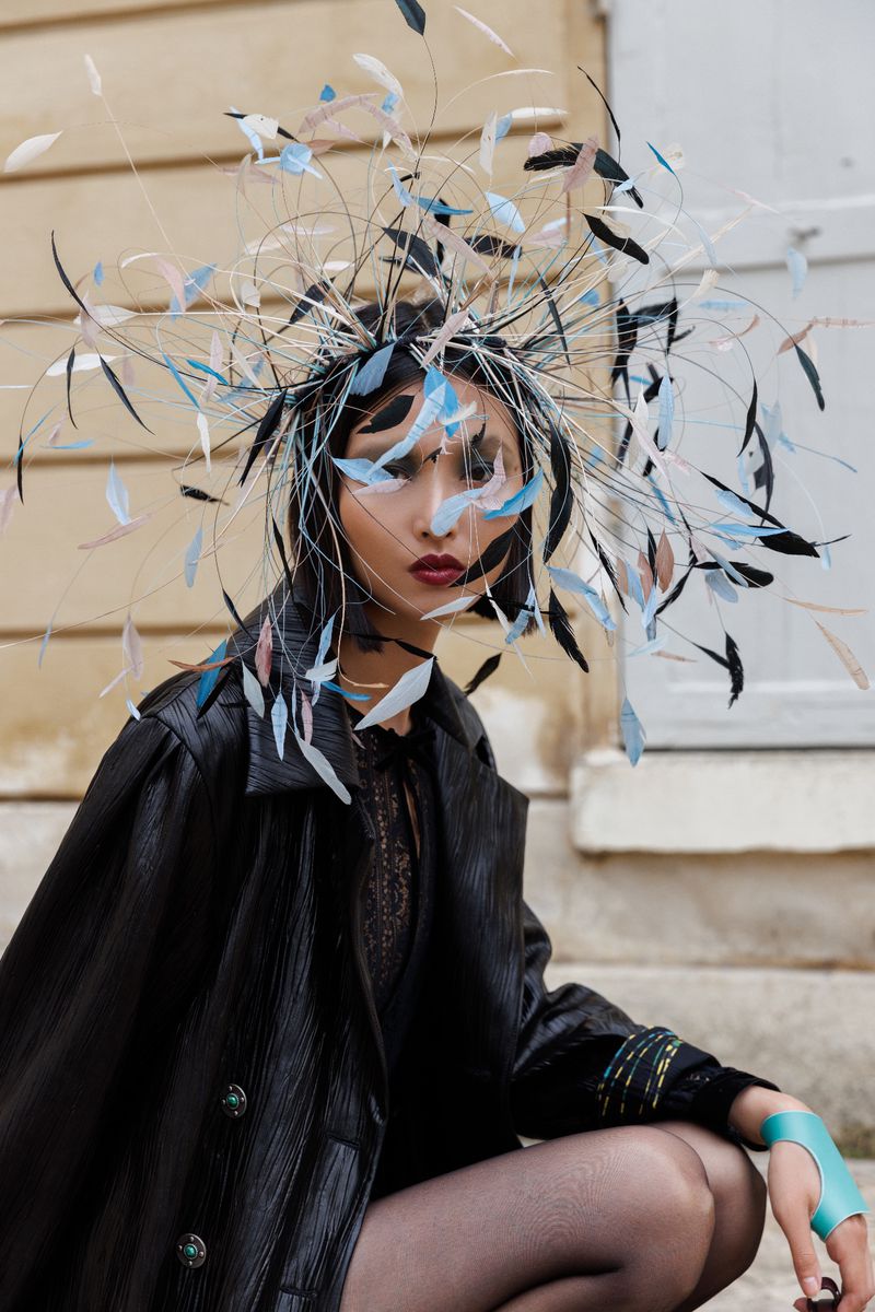 Woman in avant-garde feather headpiece and black coat poses on stone steps for a fashion editorial, showcasing creative and bold style.